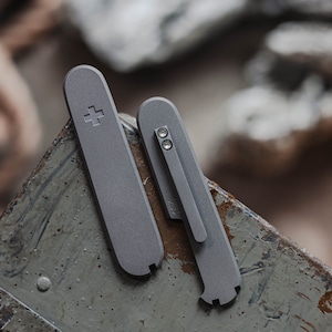 Może przedstawiać: Dwa szare metalowe narzędzia o zaokrąglonych krawędziach. Większe narzędzie ma symbol krzyża po jednej stronie. Mniejsze narzędzie ma dwa małe srebrne nity i mały otwór po boku.