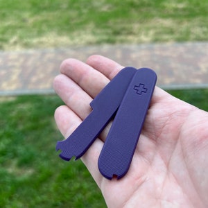 Puede incluir: Dos escamas de plástico morado para una navaja suiza. Las escamas tienen una superficie texturizada y un pequeño logotipo de cruz en una de las escamas.