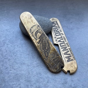 Può includere: Un coltellino tascabile in ottone con un design di Mandalorian. Il coltellino presenta un'immagine in bianco e nero di un elmetto di Mandalorian e il testo "Sono un Mandaloriano. Le armi fanno parte della mia religione."