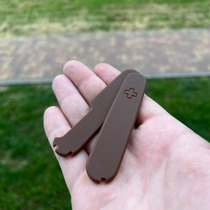 May include: Two brown plastic scales for a Swiss Army knife. The scales have a textured surface and a small cross embossed on one side.