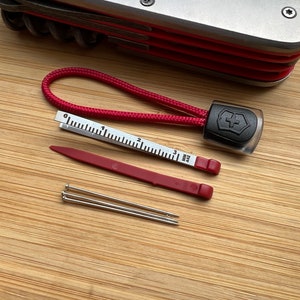 Pode incluir: Uma ferramenta multifuncional vermelha e preta com um cord&atilde;o vermelho, uma pequena r&eacute;gua, uma ferramenta de pl&aacute;stico vermelha e tr&ecirc;s pinos prateados.