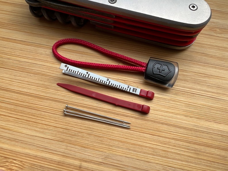 Pode incluir: Uma ferramenta multifuncional vermelha e preta com um cord&atilde;o vermelho, uma pequena r&eacute;gua, uma ferramenta de pl&aacute;stico vermelha e tr&ecirc;s pinos prateados.