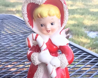 May include: A vintage porcelain figurine of a girl wearing a red hooded coat with white fur trim, holding a candy cane. She has blonde hair and a sweet expression.