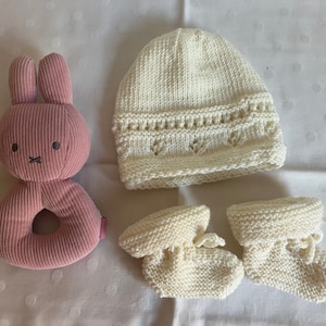 May include: A set of knitted baby clothes in white, including a hat, booties, and a pink plush bunny rattle.