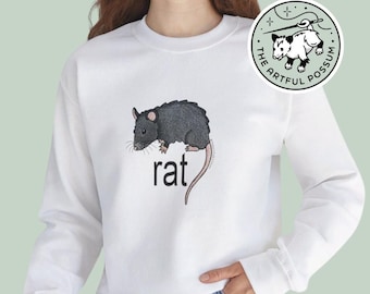 May include: White sweatshirt with a black rat illustration and the word "rat" printed on the front. The sweatshirt has a crew neck and long sleeves. The brand logo is a black and white possum with the text "The Artful Possum".