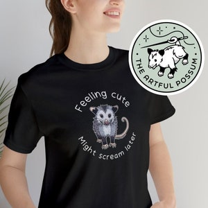 Puede incluir: Camiseta negra con el texto "Feeling cute, might scream later" que rodea un dibujo de una zarigüeya. También es visible una pegatina redonda con un dibujo de una zarigüeya y el texto "The Artful Possum".