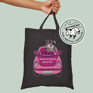 May include: Black tote bag featuring a pink car with a possum wearing sunglasses. The car has the text "the horrors persist, but so do I" and "AAAAAAA". The tote bag also has a round sticker with a possum and the text "The Artful Possum".