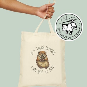May include: A cream-coloured tote bag featuring a cartoon hedgehog illustration and the text "HEY THERE DEMONS I AM NOT YA BOY." A circular sticker with a possum and needle logo is visible. The bag is held by a hand.