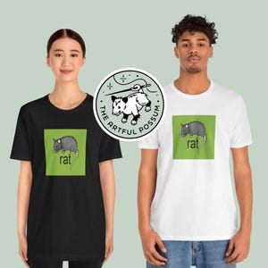 Puede incluir: Dos personas con camisetas negras y blancas con un gr&aacute;fico cuadrado verde que muestra una rata y la palabra "rat". La camiseta negra tiene una pegatina circular blanca con una ilustraci&oacute;n de zarig&uuml;eya y el texto "The Artful Possum".