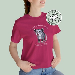 Op de afbeelding: Een roze T-shirt met een cartoon opossum-afbeelding en de tekst "I'm an acquired taste. If you don't like me, acquire some taste."