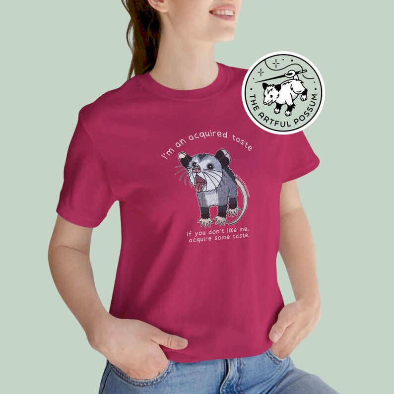 Op de afbeelding: Een roze T-shirt met een cartoon opossum-afbeelding en de tekst "I'm an acquired taste. If you don't like me, acquire some taste."