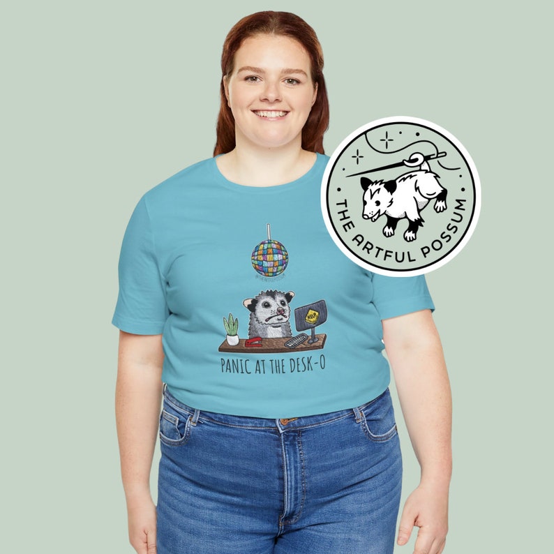 以下が含まれることがあります： ライトブルーのTシャツを着た女性。Tシャツには、コンピューターデスクに座るオポッサムのグラフィックが描かれており、その上にディスコボールがぶら下がっています。Tシャツの文字は「Panic at the Desk-O」。Tシャツの右下には、「The Artful Possum」という小さなロゴがあります。