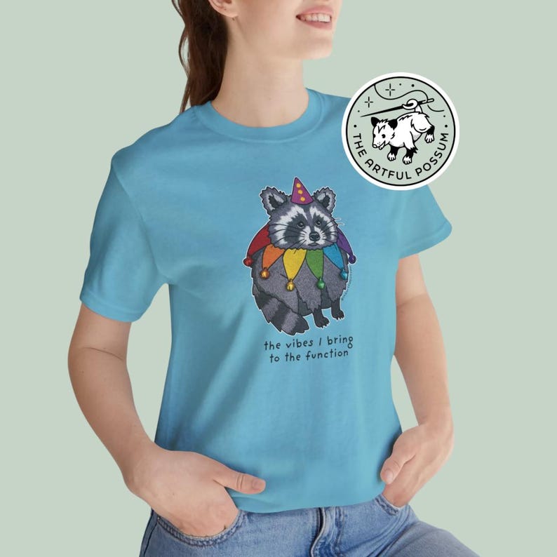 May include: A light blue t-shirt with a cartoon raccoon wearing a rainbow jester hat and the text "the vibes I bring to the function".