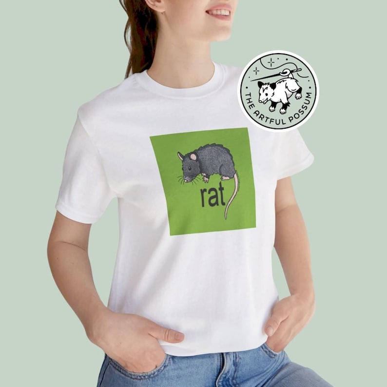 Puede incluir: Camiseta blanca con un cuadrado verde que presenta una ilustraci&oacute;n de rata negra y la palabra "rat" en texto negro. La camiseta tiene una peque&ntilde;a pegatina redonda blanca con una ilustraci&oacute;n en blanco y negro de una zarig&uuml;eya y el texto "The Artful Possum".
