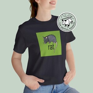 Puede incluir: Una camiseta negra con un cuadrado verde que presenta una ilustraci&oacute;n de rata negra y la palabra "rat" en texto blanco. La camiseta tiene un peque&ntilde;o logotipo circular blanco con una ilustraci&oacute;n de zarig&uuml;eya negra y el texto "The Artful Possum".