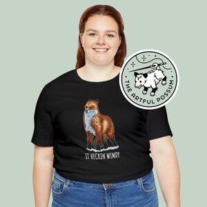 May include: A black t-shirt with a red fox illustration and the text "IT HECKIN WINDY". A sticker with a white possum illustration and the text "THE ARTFUL POSSUM" is shown on the right side of the image.