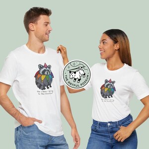 May include: Two white t-shirts with a cartoon raccoon wearing a jester hat and rainbow colors. The text on the shirts reads "The vibes I bring to the function". The shirts are being modeled by two people. A sticker with a possum and the text "The Artful Possum" is visible in the background.