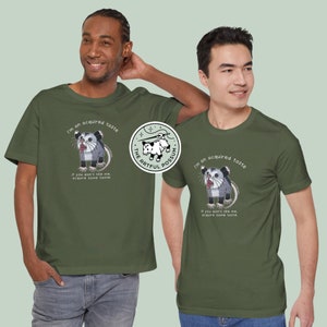 Op de afbeelding: Twee mannen dragen groene T-shirts met een afbeelding van een opossum en de tekst "I'm an acquired taste. If you don't like me, acquire some taste." De afbeelding bevat ook de tekst "The Artful Possum".