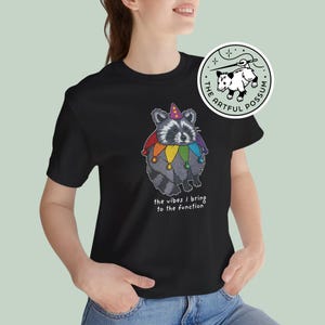 May include: Black t-shirt featuring a cartoon raccoon wearing a rainbow jester hat and the text "the vibes / bring to the function". The shirt also has a small logo with a possum in a circle.
