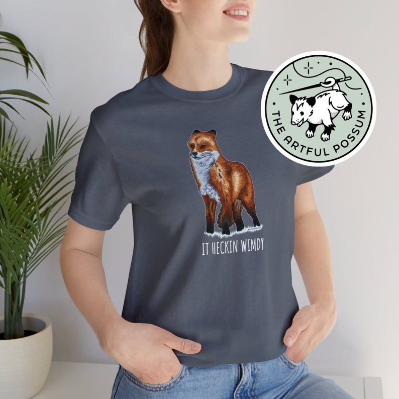 May include: A dark blue t-shirt with a red fox illustration and the text "It heckin wimdy". The t-shirt has a round, green logo with a white possum illustration and the text "The Artful Possum".