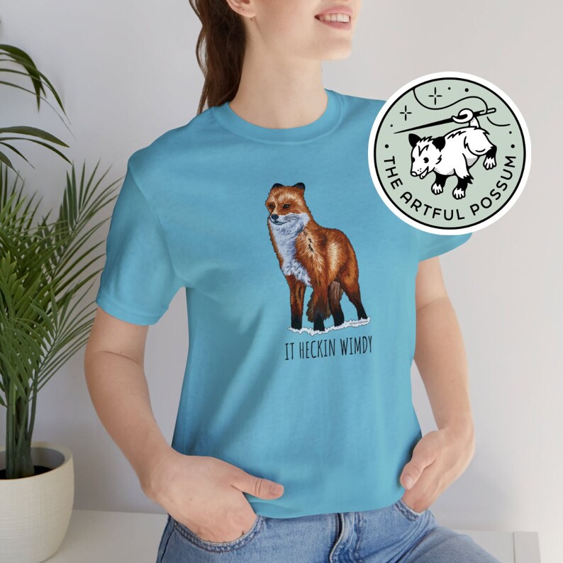 May include: A turquoise blue t-shirt with a graphic of a red fox standing on a snowy patch. The text "IT HECKIN WIMDY" is printed below the fox. A small circular patch with a possum graphic and the text "THE ARTFUL POSSUM" is visible in the background.