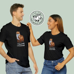 May include: Two black t-shirts with a graphic of a fox and the text "It heckin' windy". The t-shirts are being modeled by two people. A logo for "The Artful Possum" is in the background.