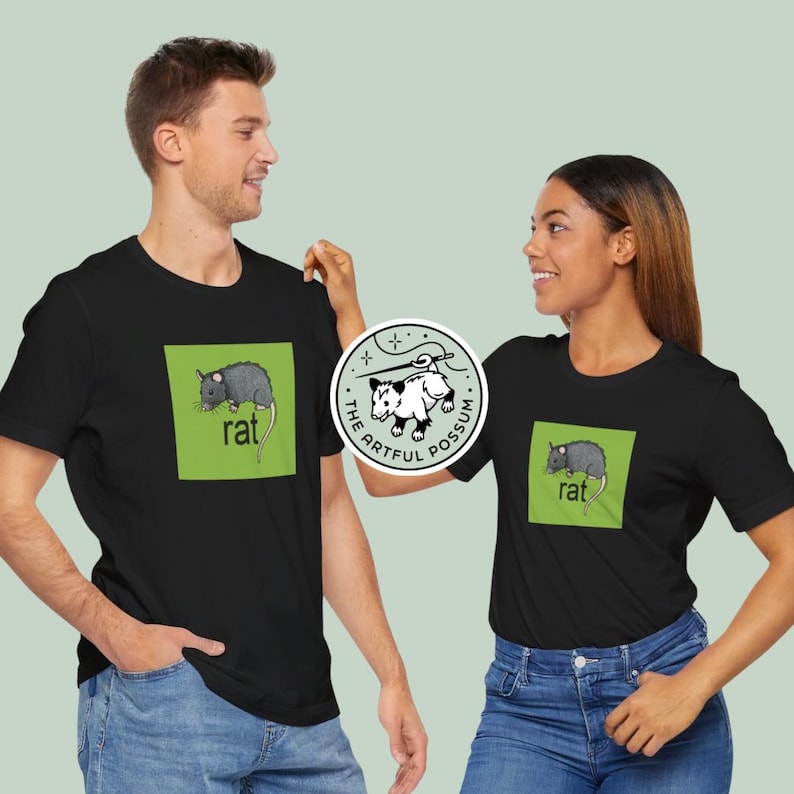 Puede incluir: Dos personas con camisetas negras con un gr&aacute;fico cuadrado verde de una rata y la palabra "rat" impresa en el cuadrado. La persona de la izquierda lleva pantalones vaqueros azules. La persona de la derecha lleva pantalones vaqueros azules y tiene una pegatina en su camiseta que dice "The Artful Possum".