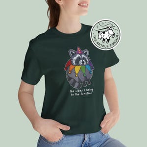 May include: A dark green t-shirt with a cartoon raccoon wearing a rainbow jester hat and the text "the vibes I bring to the function". The shirt also features a small logo with a possum and the text "The Artful Possum".