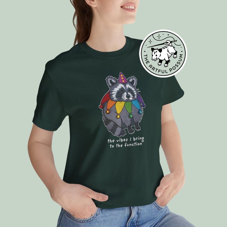 May include: A dark green t-shirt with a cartoon raccoon wearing a rainbow jester hat and the text "the vibes I bring to the function". The shirt also features a small logo with a possum and the text "The Artful Possum".