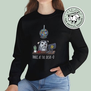 Puede incluir: Sudadera negra con un gráfico de un opossum sentado en un escritorio con una computadora, una planta, una grapadora y una bola de discoteca arriba. El texto "Panic at the Desk-O" está debajo del gráfico. El texto "The Artful Possum" está en un círculo arriba del gráfico.