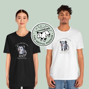 Op de afbeelding: Twee mensen dragen zwarte en witte T-shirts met een opossum-afbeelding en de tekst "I'm an acquired taste. If you don't like me, acquire some taste." De opossum-afbeelding staat ook op een aparte sticker met de tekst "The Artful Possum."