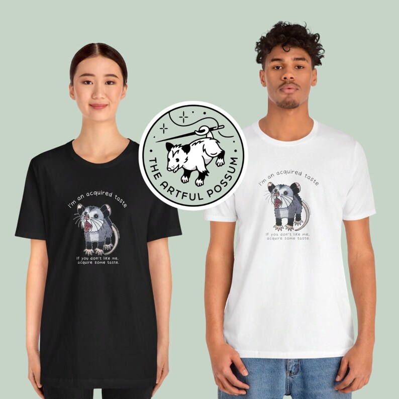 Op de afbeelding: Twee mensen dragen zwarte en witte T-shirts met een opossum-afbeelding en de tekst "I'm an acquired taste. If you don't like me, acquire some taste." De opossum-afbeelding staat ook op een aparte sticker met de tekst "The Artful Possum."