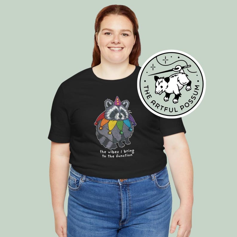 May include: A black t-shirt with a graphic of a raccoon wearing a jester hat and a rainbow collar. The text "the vibes I bring to the function" is printed below the graphic. The t-shirt is being modeled by a person. A white sticker with a black and white illustration of a possum is visible on the right side of the image.