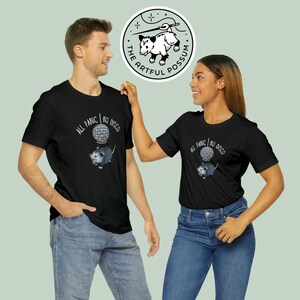 K&ouml;nnte beinhalten: Zwei Personen tragen schwarze T-Shirts mit einem Opossum-Motiv und dem Text "All Panic | No Disco". Das Opossum h&auml;lt eine Discokugel. Die T-Shirts sind auf wei&szlig;em Hintergrund.