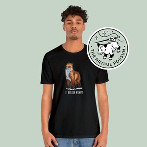 May include: Black t-shirt with a graphic of a red fox standing in the snow. The text "It heccin windy" is printed below the fox. The t-shirt is being modeled by a person. A sticker with a possum graphic and the text "The Artful Possum" is shown in the upper right corner of the image.