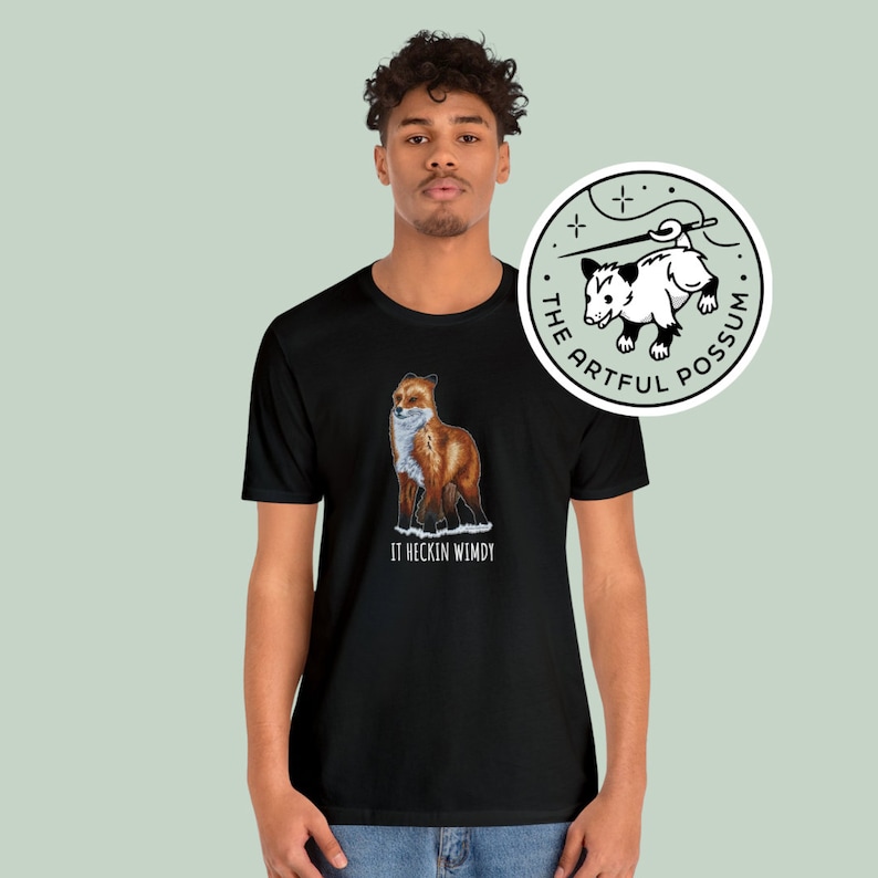 May include: Black t-shirt with a graphic of a red fox standing in the snow. The text "It heccin windy" is printed below the fox. The t-shirt is being modeled by a person. A sticker with a possum graphic and the text "The Artful Possum" is shown in the upper right corner of the image.