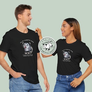 Op de afbeelding: Twee mensen dragen zwarte T-shirts met een opossum-afbeelding en de tekst "I'm an acquired taste. If you don't like me, acquire some taste." en "The Artful Possum".