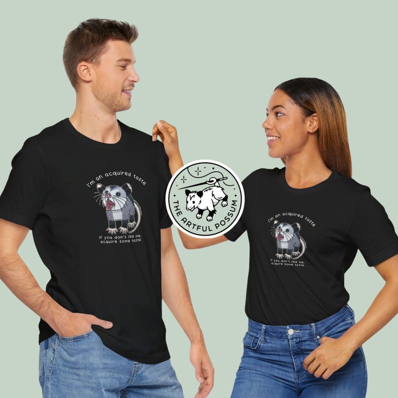 Op de afbeelding: Twee mensen dragen zwarte T-shirts met een opossum-afbeelding en de tekst "I'm an acquired taste. If you don't like me, acquire some taste." en "The Artful Possum".