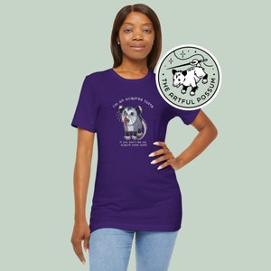 Op de afbeelding: Paars T-shirt met een opossum-afbeelding en de tekst "I'm an acquired taste. If you don't like me, acquire some taste." De tekst "The Artful Possum" staat in een cirkel met een naald en draad.