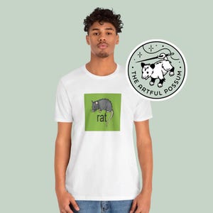Puede incluir: Camiseta blanca con un cuadrado verde que presenta una ilustraci&oacute;n de rata negra y gris y la palabra "rat" en texto negro. La camiseta tiene una pegatina redonda blanca con una ilustraci&oacute;n de zarig&uuml;eya negra y blanca y el texto "The Artful Possum" en texto negro.