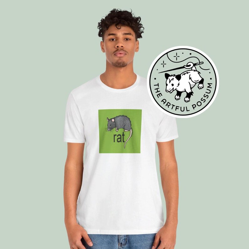 Puede incluir: Camiseta blanca con un cuadrado verde que presenta una ilustraci&oacute;n de rata negra y gris y la palabra "rat" en texto negro. La camiseta tiene una pegatina redonda blanca con una ilustraci&oacute;n de zarig&uuml;eya negra y blanca y el texto "The Artful Possum" en texto negro.