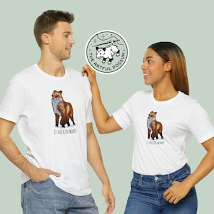May include: Two white t-shirts with a graphic of a fox and the text "It heckin' windy". The t-shirts are being modeled by two people. A green circle with a possum graphic and the text "The Artful Possum" is in the background.