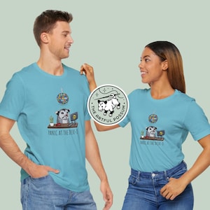 以下が含まれることがあります： 2人の人が、薄い青色のTシャツを着ています。Tシャツには、オポッサムのグラフィックと「Panic at the desk-o」というテキスト、そしてディスコボールのグラフィックが描かれています。オポッサムのグラフィックはヘッドセットを装着し、コンピューターのあるデスクに座っています。Tシャツには、「The Artful Possum」というテキストのロゴも付いています。