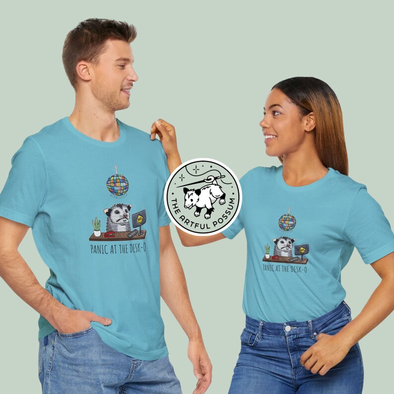 以下が含まれることがあります： 2人の人が、薄い青色のTシャツを着ています。Tシャツには、オポッサムのグラフィックと「Panic at the desk-o」というテキスト、そしてディスコボールのグラフィックが描かれています。オポッサムのグラフィックはヘッドセットを装着し、コンピューターのあるデスクに座っています。Tシャツには、「The Artful Possum」というテキストのロゴも付いています。