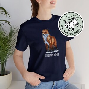 May include: A navy blue t-shirt with a graphic of a red fox and the text "IT HECKIN WIMDY". The t-shirt is being worn by a person. A sticker with a possum and the text "THE ARTFUL POSSUM" is visible in the background.