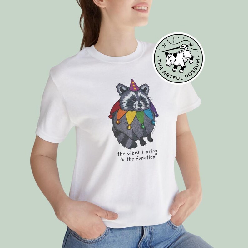 May include: White t-shirt with a graphic of a raccoon wearing a jester hat and rainbow-colored jester bells. The text "the vibes I bring to the function" is printed below the raccoon. The The Artful Possum logo is in the top right corner.