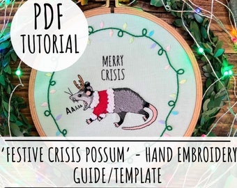 Peut inclure: Un tutoriel PDF pour un projet de broderie à la main représentant un opossum portant un chapeau de Père Noël et des bois de renne. L'opossum est brodé sur un tissu bleu clair avec le texte "Merry Crisis" au-dessus. La broderie est encadrée dans un cercle en bois.