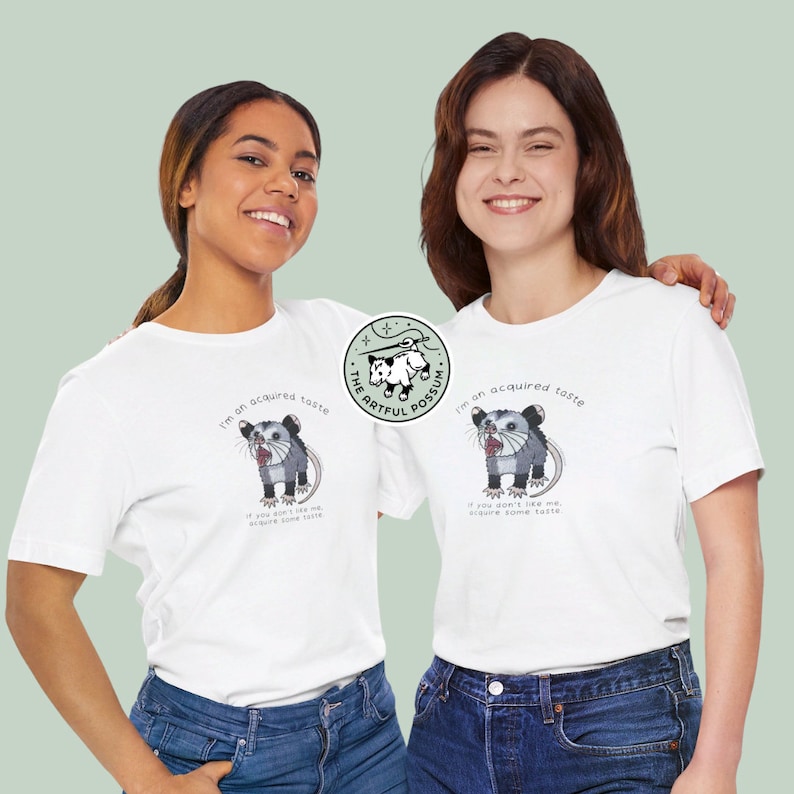 Op de afbeelding: Twee mensen dragen witte T-shirts met een afbeelding van een opossum en de tekst "I'm an acquired taste. If you don't like me, acquire some taste." De tekst is in zwart. De opossum is een cartoonillustratie met een wit lichaam en zwarte ogen. De T-shirts zijn wit en de mensen dragen blauwe jeans.