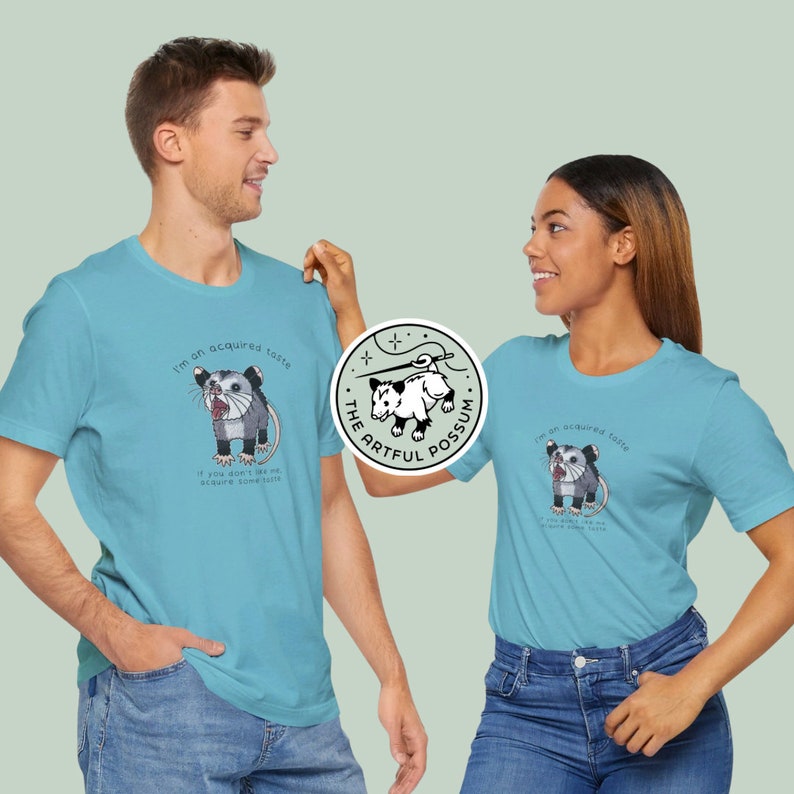 Op de afbeelding: Twee mensen dragen lichtblauwe T-shirts met een opossum-afbeelding en de tekst "I'm an acquired taste. If you don't like me acquire some taste." en "The Artful Possum".