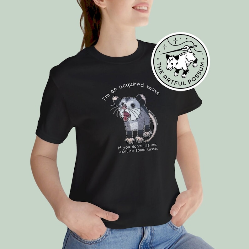Op de afbeelding: Zwart T-shirt met een witte afbeelding van een opossum met de tekst "I'm an acquired taste" en "If you don't like me, acquire some taste." De tekst "THE ARTFUL POSSUM" staat in een cirkel met een ster en een naald.